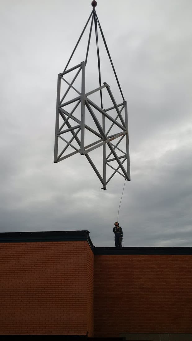 A steel truss lifted onto a rooftop by crane, with an ironworker guiding it in
